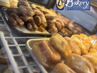 Display case with trays of glazed donuts and pastries, with a blue and yellow &quot;Ole Country Bakery&quot; sign featuring a woman in traditional dress in the top right corner.