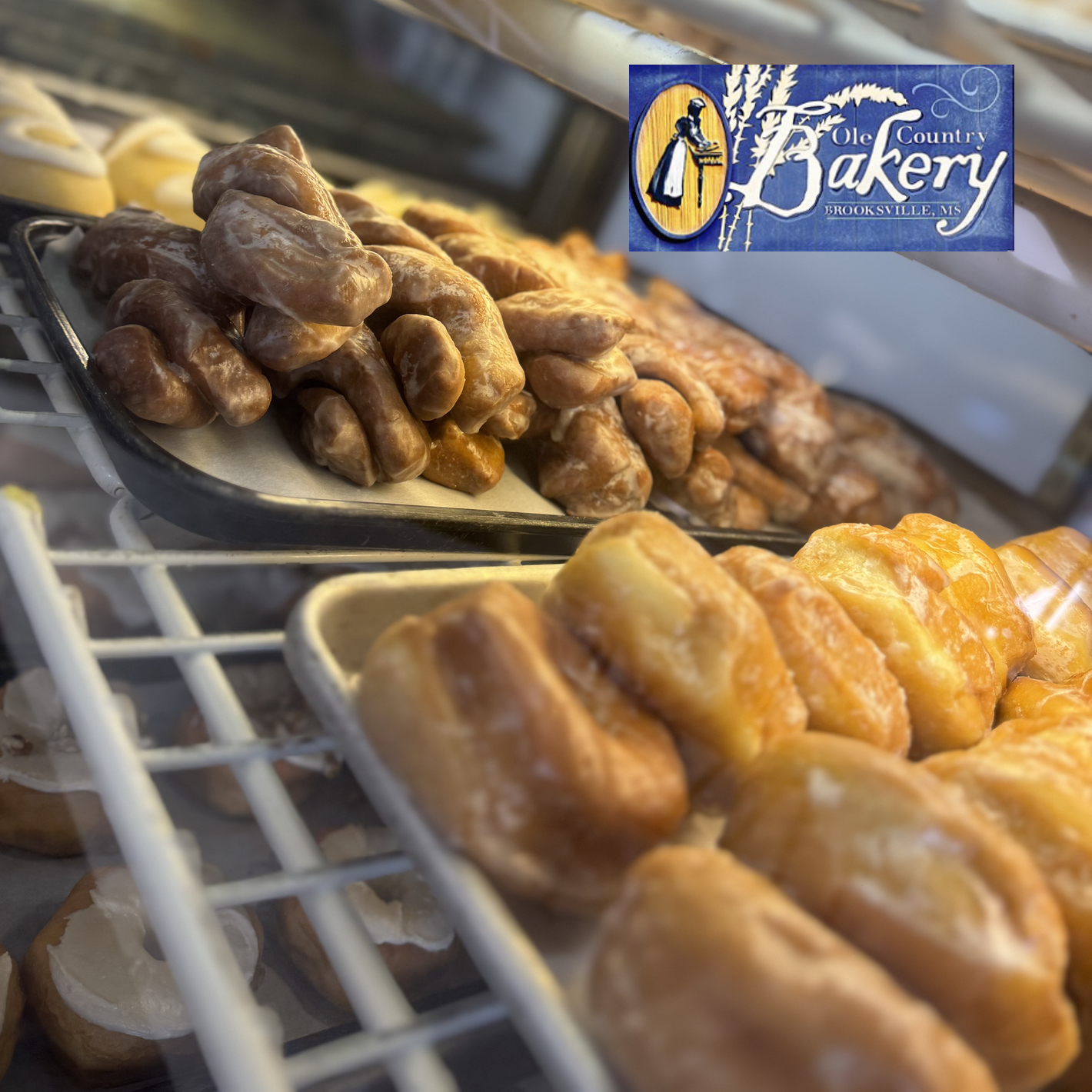Display case with trays of glazed donuts and pastries, with a blue and yellow "Ole Country Bakery" sign featuring a woman in traditional dress in the top right corner.