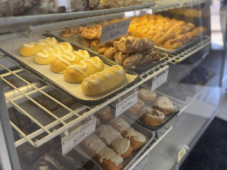A display case filled with assorted pastries and donuts, including eclairs, apple fritters, and iced cake donuts, arranged on metal trays.