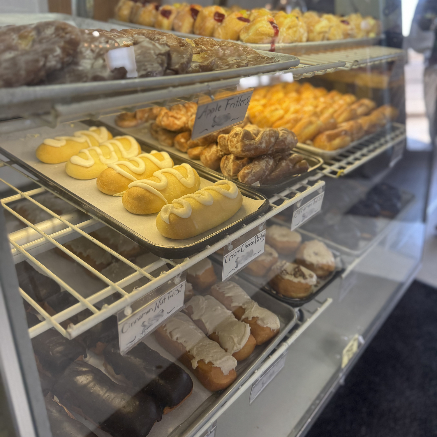 A display case filled with assorted pastries and donuts, including eclairs, apple fritters, and iced cake donuts, arranged on metal trays.