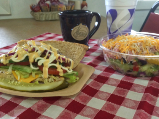A sandwich with lettuce, tomato, cheese, and sauce from the old country bakery is served with a pickle, a salad with shredded cheese, a mug, and a soft drink on a red and white checkered tablecloth.