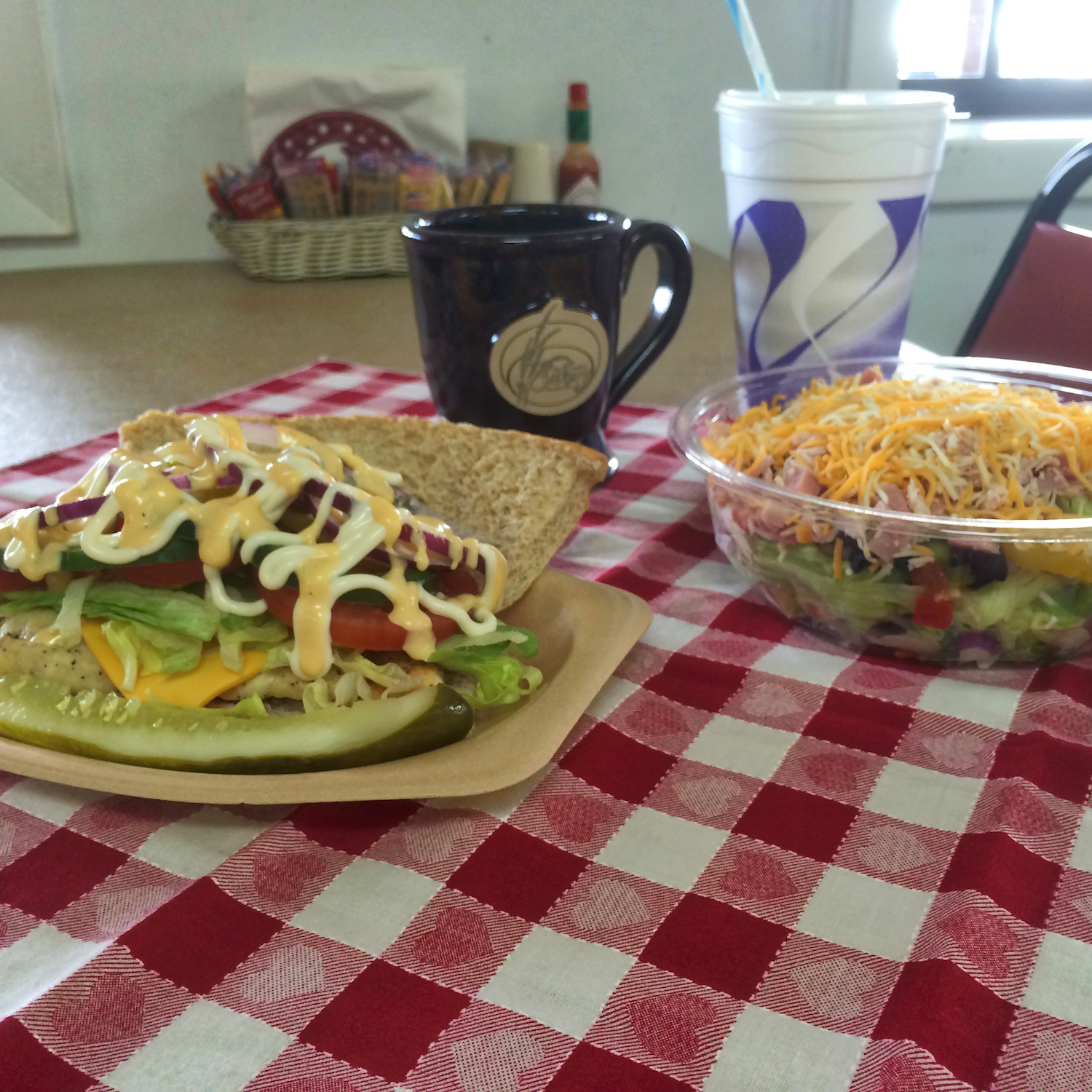 A sandwich with lettuce, tomato, cheese, and sauce from the old country bakery is served with a pickle, a salad with shredded cheese, a mug, and a soft drink on a red and white checkered tablecloth.